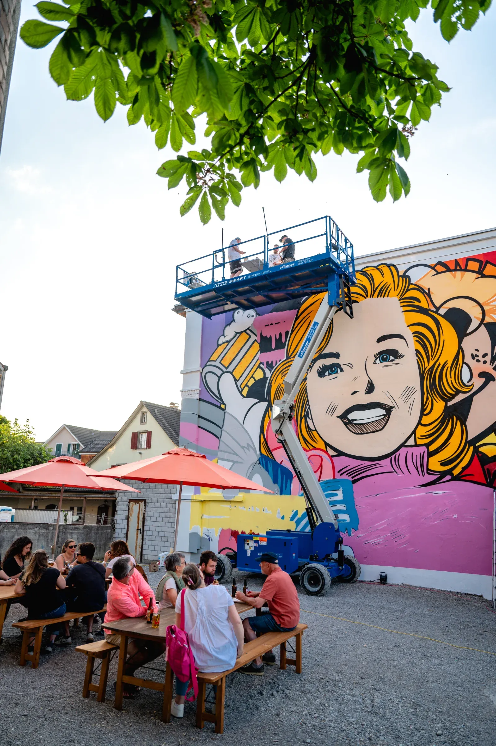 Three people work from a lift platform on BustArt's mural, guests sit at beer tables below