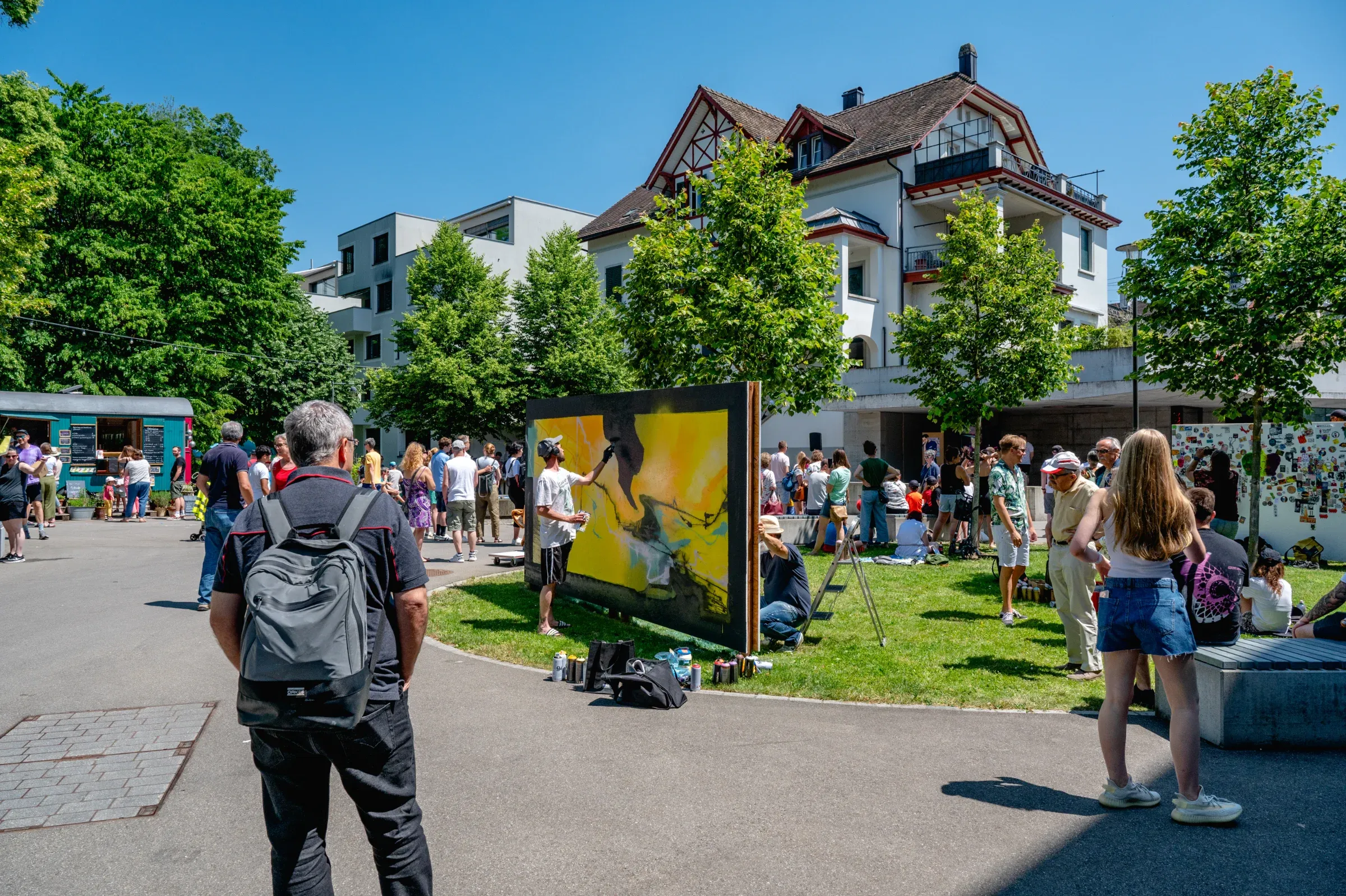 A sprayer paints a large yellow panel in Lindenpark, onlookers standing around on the lawn