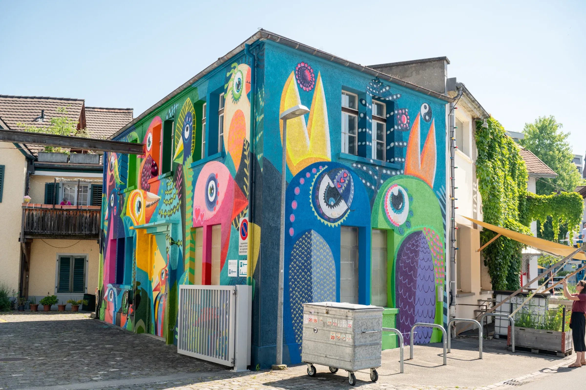A corner house painted all round with colourful birds, bike racks and a bin in front