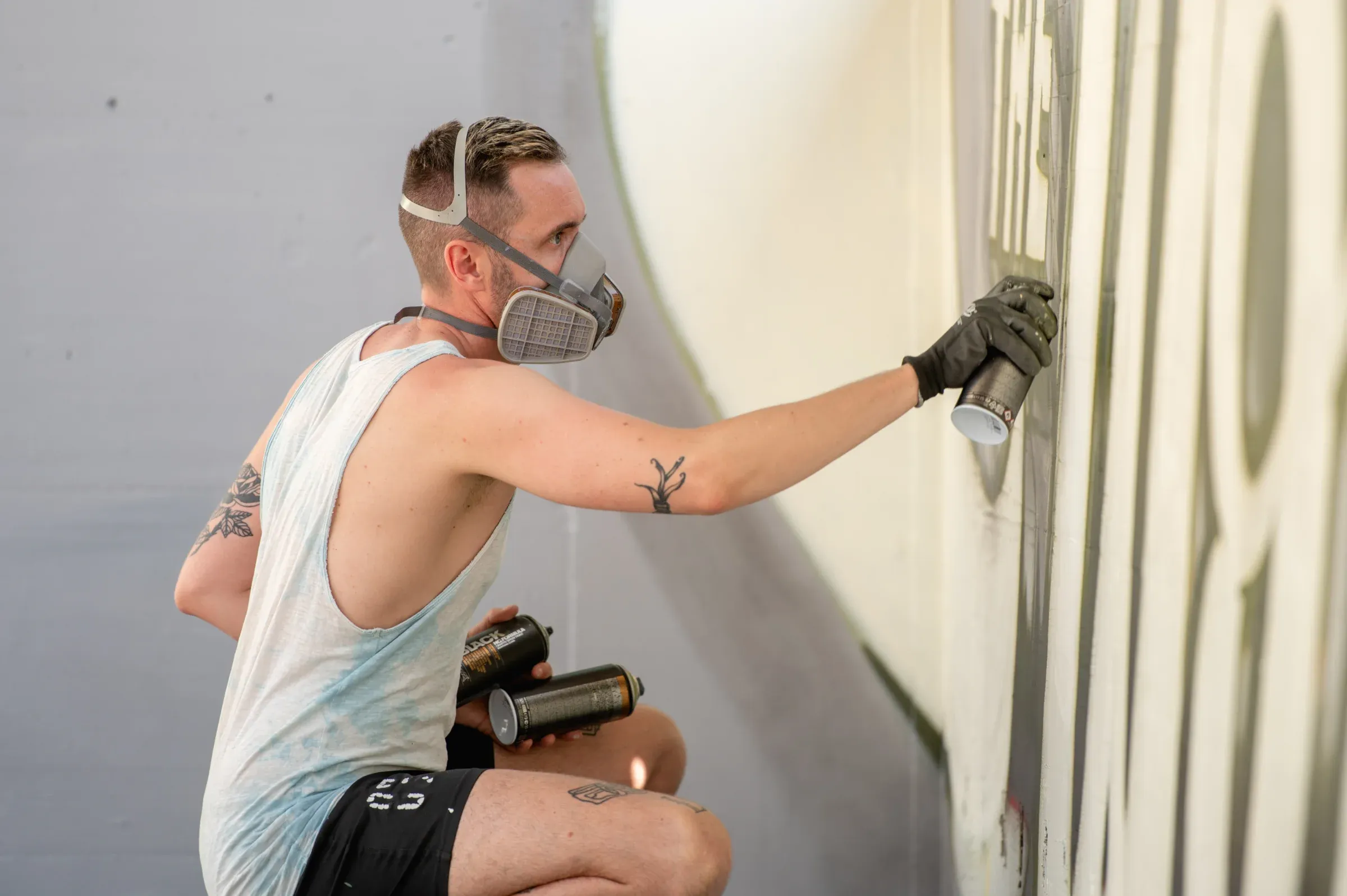 A sprayer wearing a respirator kneels in front of a pale wall, cans beside him