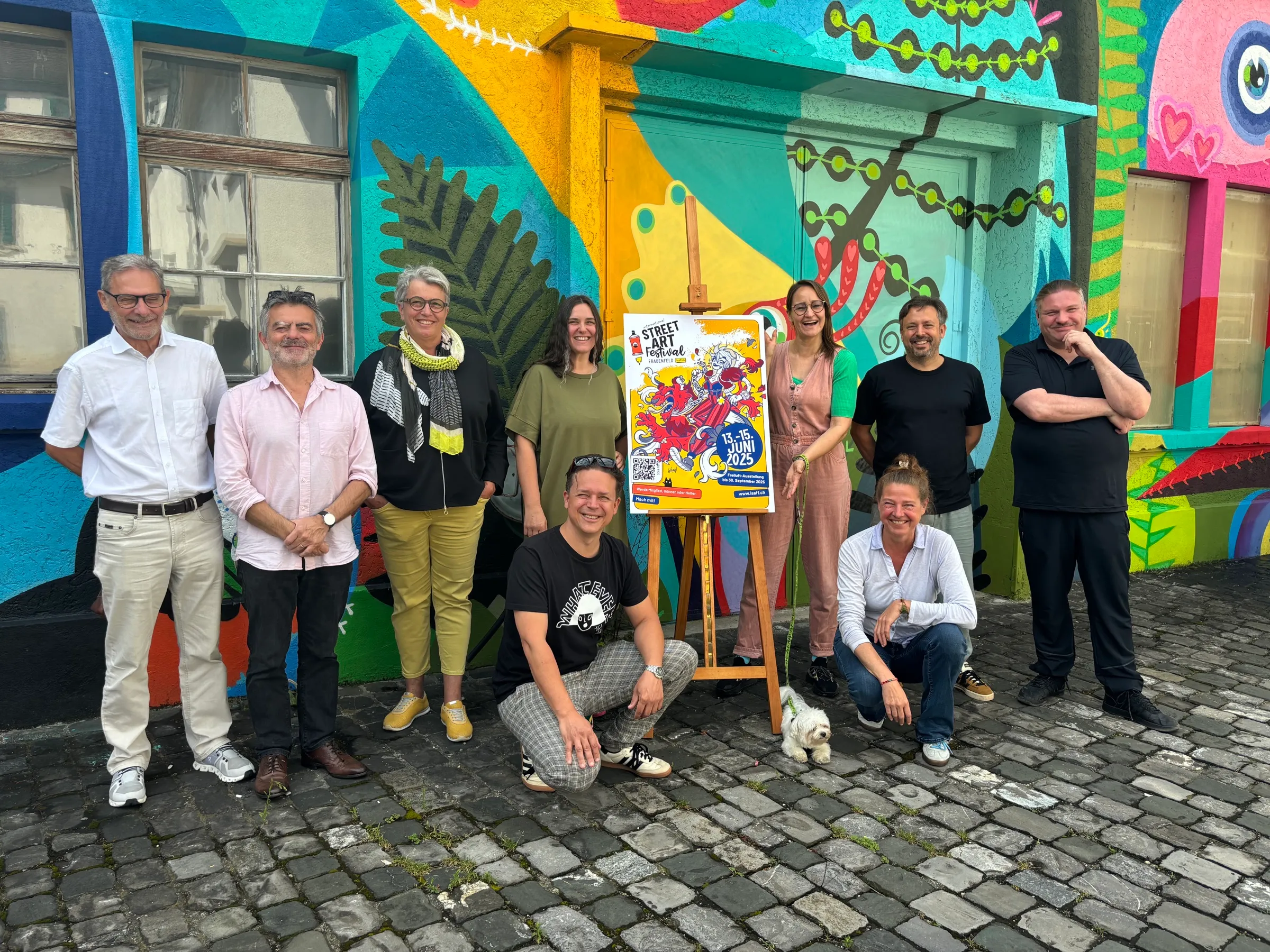 Vorstand und Geschäftsstelle von Pro Streetart Frauenfeld vor einer bemalten Wand, daneben das Festivalplakat auf einer Staffelei